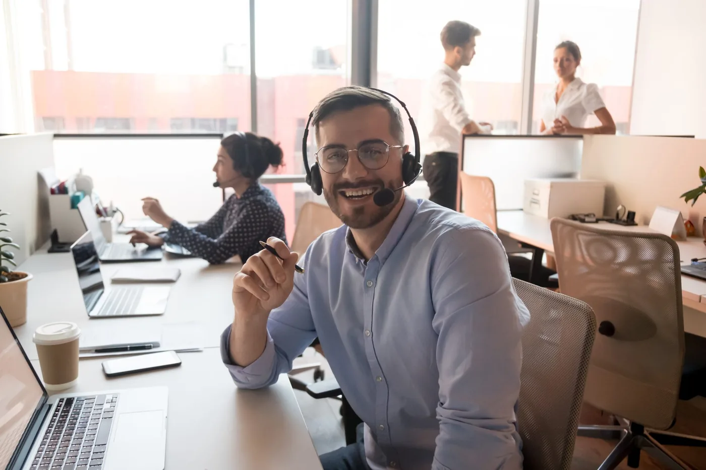 Servicedeskmedewerker met een headset en een pen in de hand aan een bureau met laptops