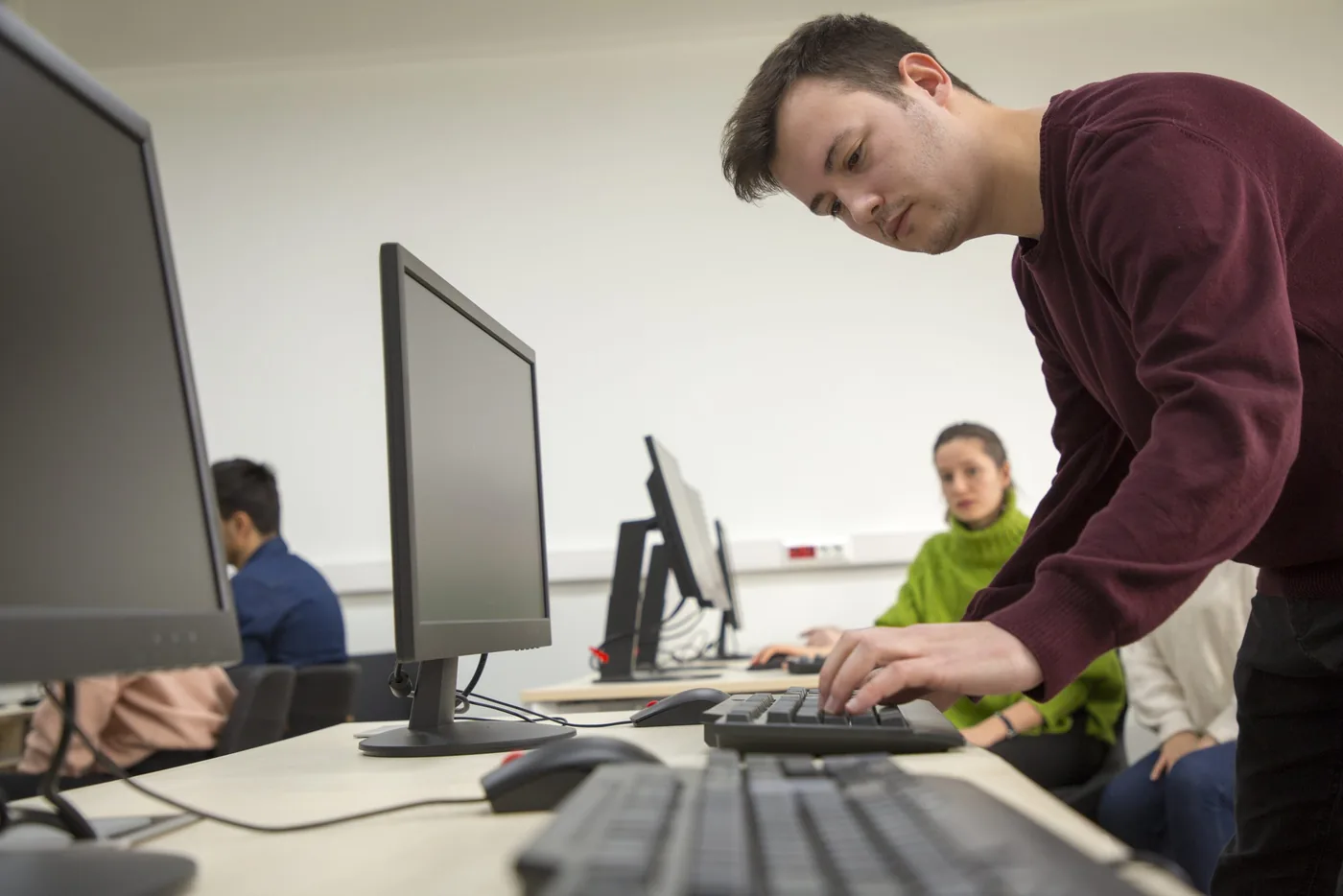 Iemand die over een bureau leunt en typt in een lokaal met meerdere computeropstellingen